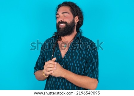Dreamy Caucasian man with beard wearing blue shirt over blue background with pleasant expression, closes eyes, keeps hands crossed near face, thinks about something pleasant