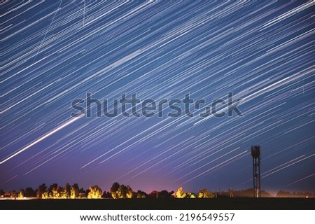 Amazing Unusual Stars Effects In Sky. Star Lines Move In Sky Above Rural Landscape. Soft Blue-pink Colours. Water Tower In Background. Large Exposure. Star Trails On Night Sky. Imagination, Fantasy.