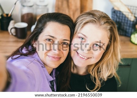 Girlfriends laugh merrily and take selfies in the kitchen. Two girls are photographed on the phone. Brunette and blonde have fun. Women looking at smartphone screen and smiling