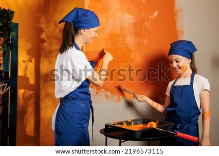 Young people painting walls with orange color paint and brush with bristles, using redecorating tools. Adult and little girl being messy and having fun renovating interior apartment space.