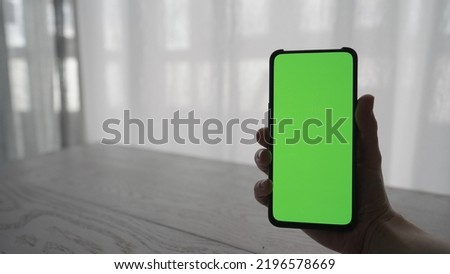 Man showing and using smarphone with green screen while sitting behind white oak table, wide photo