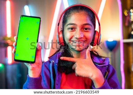Happy teenager girl kid with headset showing green screen mobile phone by pointing finger by looking camera at home - concept of app advertisement, entertainment and promotion.