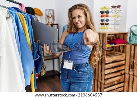 Young caucasian woman working as manager at retail boutique holding laptop pointing with finger to the camera and to you, confident gesture looking serious 
