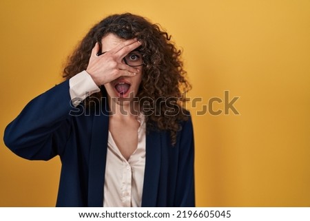 Hispanic woman with curly hair standing over yellow background peeking in shock covering face and eyes with hand, looking through fingers with embarrassed expression. 