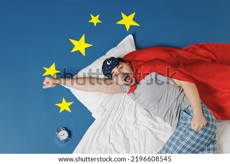 Top view shocked confused young man in pajamas jam sleep mask red superhero suit rest relax home lie make fly gesture isolated on dark blue sky background. Night bedtime supernatural abilities concept