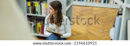 teenage girl sitting on floor with book and looking away in library, banner