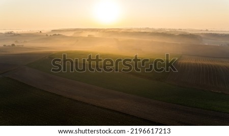 Bright yellow sunrise and glare over green and gold farm fields. Thick fog and tree silhouettes in the background. Landscape of Roztocze Poland. Horizontal shot. High quality photo