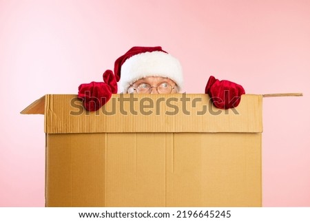 Portrait of senior man in image of Santa Claus hiding inside cardboard box. Presents, gifts for holidays. Delivery service. Concept of gifts, shopping, emotions, facial expression. Unpacking box.