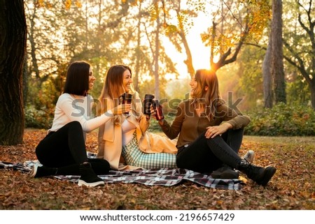 Group of three female friends smiling, sitting on picnic blanket in the park and drinking coffee, having a good time, fall season  Royalty-Free Stock Photo #2196657429