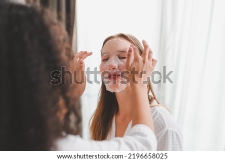 Beautiful young Caucasian woman in white bathrobe applying a revitalising mask her friend's face. Young happy Caucasian girls doing facial skin beauty treatment with friends at home.