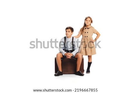 Portrait of stylish children in retro styled outfits posing, sitting on retro suitcase isolated on white studio background. Travelling. Concept of childhood, friendship, fun, lifestyle, fashion