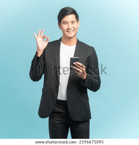 Good looking businessman portrait using smartphone and showing okay sign isolated on blue background