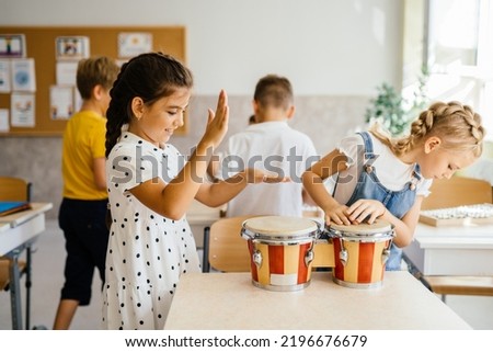 Group of excited children in the music class. Two cute little pupils girls with braids play with barabans at classroom. Back to school concept. Royalty-Free Stock Photo #2196676679