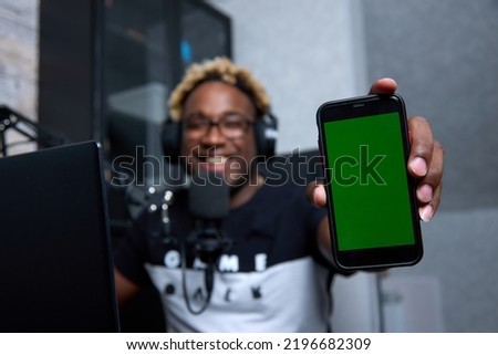 Happy African man holds a smartphone and demonstrates the good ones on his screen while sitting at laptop. A dark-skinned guy with glasses recommends installing app on the phone, smiling at the camera
