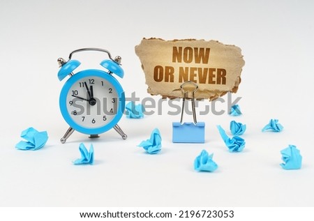 Time concept. On a white surface, an alarm clock, an office clip, crumpled paper and a cardboard sign with the text - Now or Never