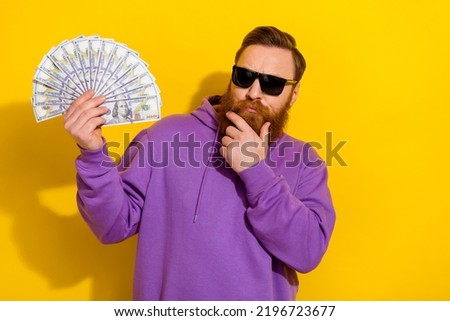 Photo of strict unsure man wear violet sweatshirt arm chin looking cash fan isolated yellow color background