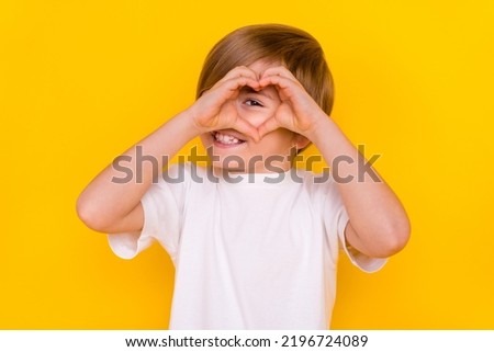 Portrait of handsome funny cheerful pre-teen boy looking through heart symbol isolated over bright yellow color background