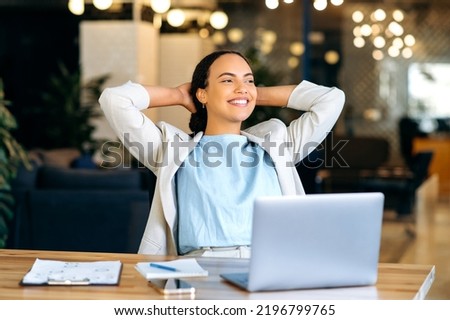 Calm relaxed mixed race female employee resting during work time, sits in modern office, puts hands behind head, feels satisfied by project done, job promotion, looking in distance, smiling friendly Royalty-Free Stock Photo #2196799765