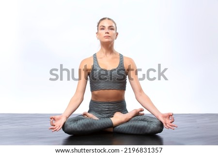 Young woman in gray, two-piece yoga outfit, seated on a black floor in a lotus yoga pose against a white background.