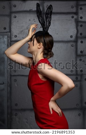 Young woman in black rabbit or hare fancy mask and red dress. Hand is on forehead. In the background there is a metal wall with rivets . Royalty-Free Stock Photo #2196891353