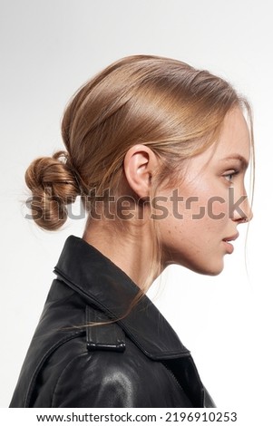 Cropped close-up shot of a blonde young woman with a chignon. The woman in the black jacket with the updo is on a white background. Side view.