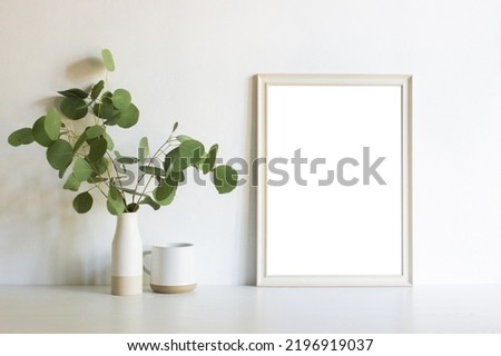 White blank frame with green leaves and coffee cups over the white wall. 