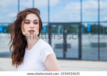 Happy young model makes a close-up photo. Her hair is blowing in the wind. Photo on the street. Delighted young lady with long dark hair in a t-shirt smiles happily and looks at the camera.