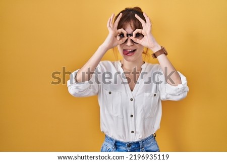 Young beautiful woman wearing casual shirt over yellow background doing ok gesture like binoculars sticking tongue out, eyes looking through fingers. crazy expression. 