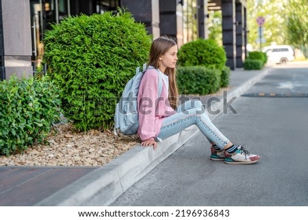A beautiful teenage girl with a backpack is sitting on the road  before school. The concept of returning to school, education and childhood