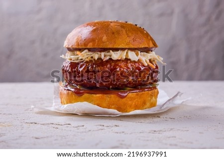 Hamburger with beef cutlet in a sauce with cabbage, ketchup and pickles that stands on white paper on a rough concrete table and against the background of a concrete wall
