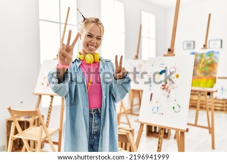 Young caucasian girl at art studio smiling with tongue out showing fingers of both hands doing victory sign. number two. 