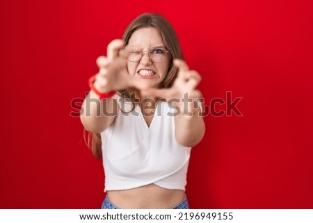 Young caucasian woman standing over red background shouting frustrated with rage, hands trying to strangle, yelling mad 