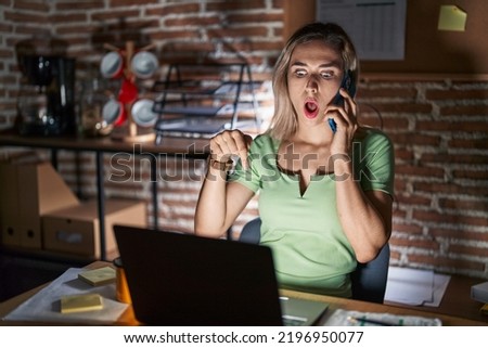 Young beautiful woman working at the office at night speaking on the phone pointing down with fingers showing advertisement, surprised face and open mouth 