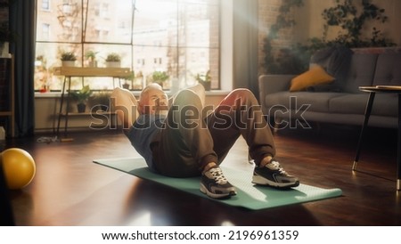 Senior Man Exercising and Training at Home on Sunny Morning. Middle Aged Male Doing Crunches, Stretching Back, Body and Muscles in Loft Stylish Apartment Living Room for Wellness and Fitness.