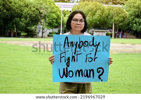 Asian middle aged female shows cardboard which has texts'Anybody Fight for Woman?' in the middle of grass lawn, concept for protesting and calling all people to support and encourage woman's power.