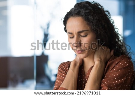 Hispanic woman overtired working in modern office businesswoman has severe neck pain, massages neck muscles, business woman in casual clothes and curly hair. Royalty-Free Stock Photo #2196971011