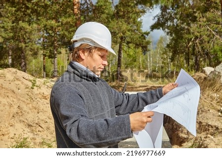 Engineer with a drawing on the construction site. A man in a white helmet. The engineer looks at the drawing. Royalty-Free Stock Photo #2196977669
