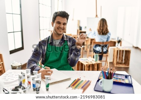 Young artist man at art studio with a big smile on face, pointing with hand finger to the side looking at the camera. 