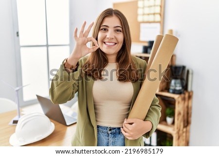 Young brunette woman holding paper blueprints at the office doing ok sign with fingers, smiling friendly gesturing excellent symbol 