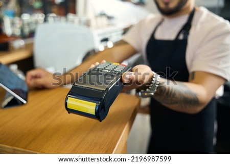 Young tattooed barista extending hand with a payment terminal