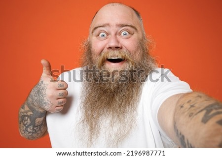 Close up fat pudge obese chubby overweight tattooed blue-eyed bearded man in white t-shirt do selfie shot on mobile phone show thumb up like gesture isolated on red orange background studio portrait