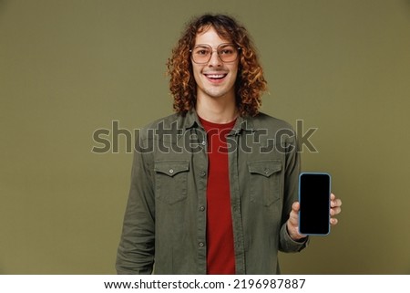 Smiling young brunet curly man 20s wears khaki shirt jacket glasses hold in hand use show mobile cell phone with blank screen workspace area isolated on plain olive green background studio portrait