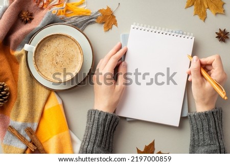 First person top view photo of woman's hands in jumper writing in notepad cup of coffee on saucer plaid fallen maple leaves pine cone anise cinnamon sticks on isolated grey background with copyspace
