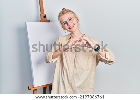 Beautiful caucasian woman with blond hair standing by painter easel stand smiling in love showing heart symbol and shape with hands. romantic concept. 
