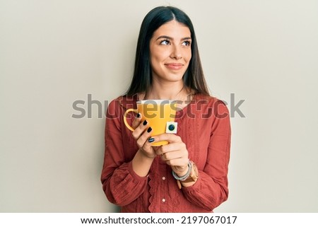 Young hispanic woman holding cup of tea smiling looking to the side and staring away thinking.  Royalty-Free Stock Photo #2197067017