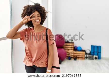 African american woman with afro hair holding yoga mat at pilates room doing peace symbol with fingers over face, smiling cheerful showing victory 