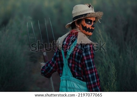 Girl with her back turned dressed as a scarecrow with halloween make-up in a green orchard with a pitchfork in her hand as she holds it and looks back at it