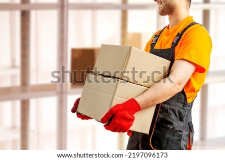 Young man holding cardboard package working in warehouse among racks and shelves. Delivery man with box. Staff laborer, orange uniform cap, t-shirt, coveralls service moving delivering orders goods.  Royalty-Free Stock Photo #2197096173