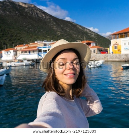 A girl in a hat smiles and takes a selfie in front of a stunning landscape with the sea, yachts and mountains on a sunny day. A woman travels to beautiful places and takes pictures on her smartphone.