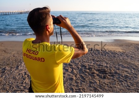 Lifeguard on the beach looking through binoculars. Safety while swimming, handsome brunette male lifeguard on the beach,back shot looking at the blue sea, copy space. Royalty-Free Stock Photo #2197145499